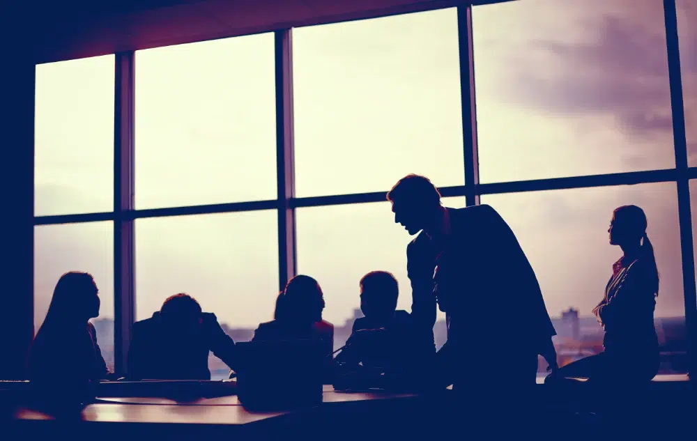 Silhouettes of a team gathered around a conference table at a proposal kickoff meeting, with a city skyline visible through large windows behind them Silhouettes of a team gathered around a conference table at a proposal kickoff meeting, with a city skyline visible through large windows behind them